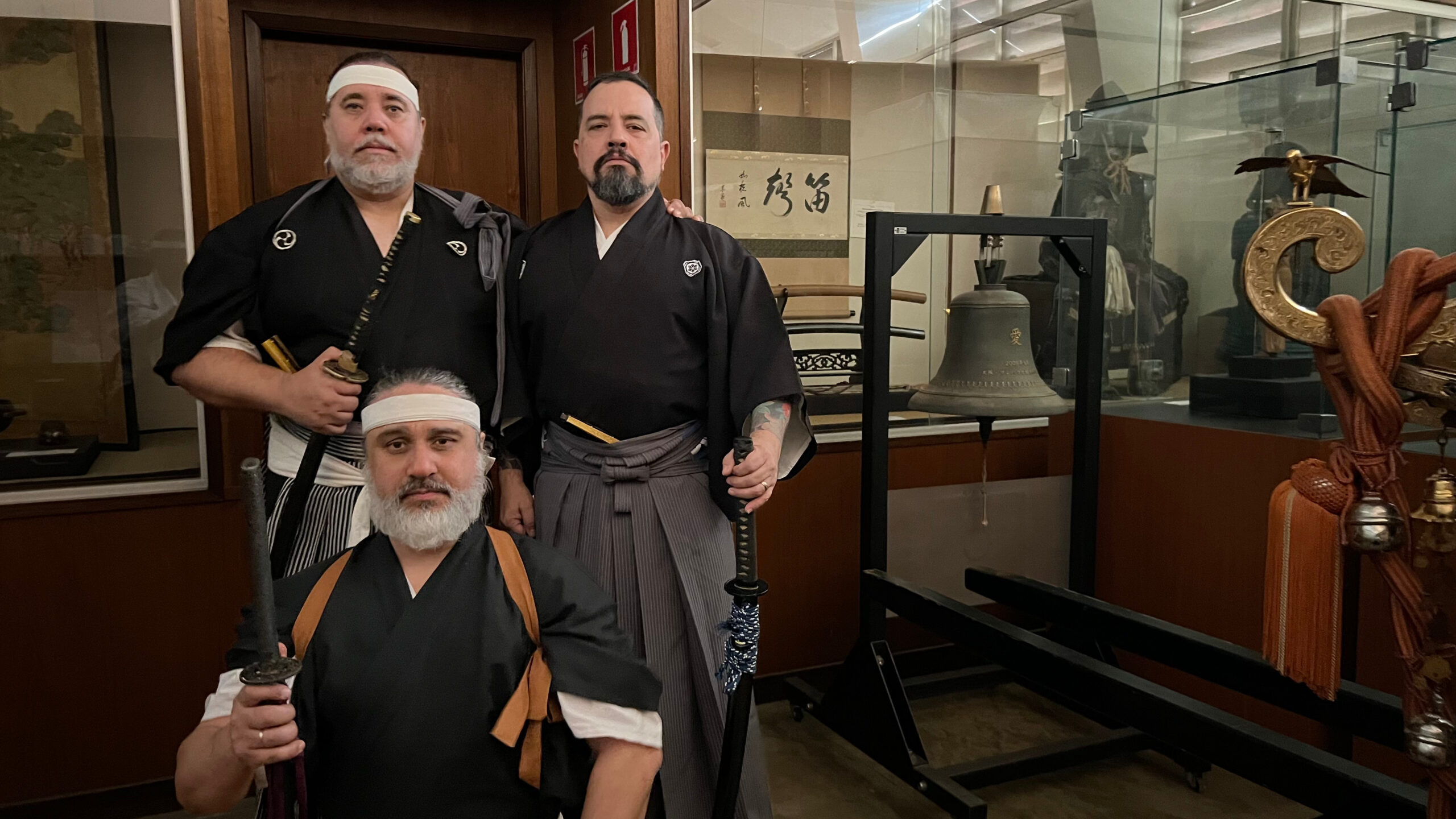 Três professores reunidos diante de um acervo de objetos japoneses
